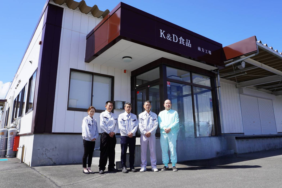 K&D食品 南方工場の外観と、正面に並ぶスタッフ